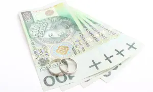 Dwa obrączki na stosie banknotów 100 zł. Symbolizuje to finansowe aspekty małżeństwa, co to jest intercyza.