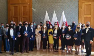 Grupa osób w garniturach i sukienkach, z flagami Polski w tle. Wśród nich radca prokuratorii generalnej, odbierający dokumenty.