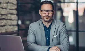 Mężczyzna w okularach i garniturze siedzi przy biurku z laptopem. To przykład osoby prawnej, która reprezentuje firmę.