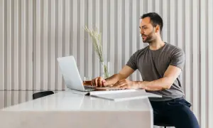 Mężczyzna w szarym t-shircie pracuje na laptopie przy białym stole.