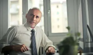 Starszy mężczyzna w białej koszuli i krawacie, z siwą brodą, siedzi przy biurku. Jego kod zawodu to pracownik administracyjny.