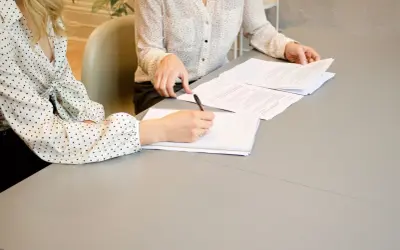 Dwie kobiety omawiają prosta umowa na wykonanie usługi. Jedna pisze w notatniku, druga przegląda dokumenty.