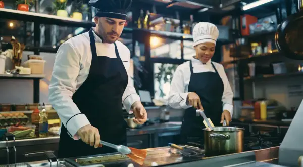 Dwóch kucharzy w kuchni restauracyjnej przygotowuje posiłek. Mężczyzna z przodu używa łopatki, kobieta z tyłu miesza coś w garnku. Kod zawodu kucharz.