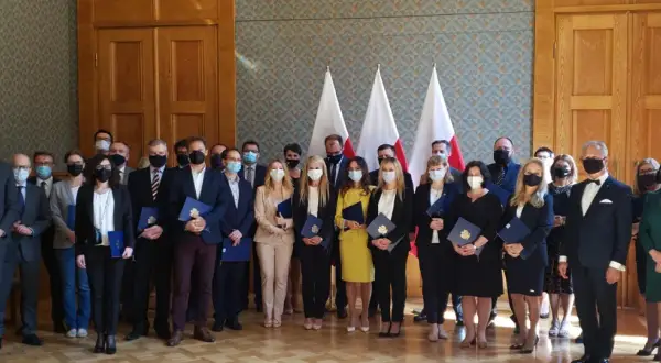 Grupa osób w garniturach i sukienkach, z flagami Polski w tle. Wśród nich radca prokuratorii generalnej, odbierający dokumenty.