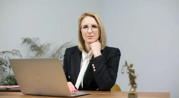 Kobieta w okularach i żakiecie siedzi przy biurku z laptopem i statuetką Temidy.