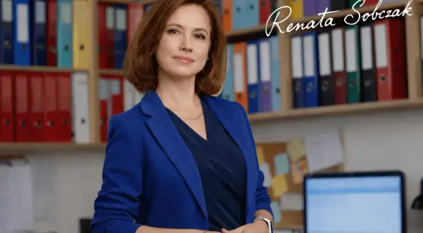 Kobieta w niebieskiej marynarce stoi przed regałami z kolorowymi segregatorami.