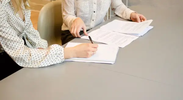 Dwie kobiety omawiają prosta umowa na wykonanie usługi. Jedna pisze w notatniku, druga przegląda dokumenty.