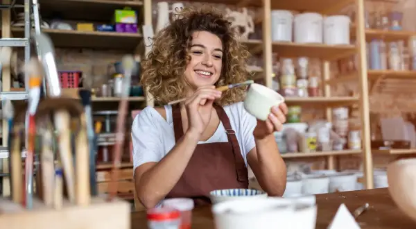 Uśmiechnięta artystka maluje ceramikę w swojej pracowni. To miejsce, gdzie powstaje i sprzedawana jest produkcja i sprzedaż rękodzieła pkd.