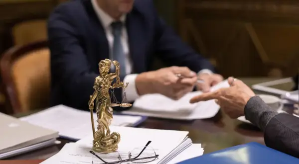 Figurka Temidy, dokumenty i okulary na stole, dwóch mężczyzn w garniturach przy biurku.
