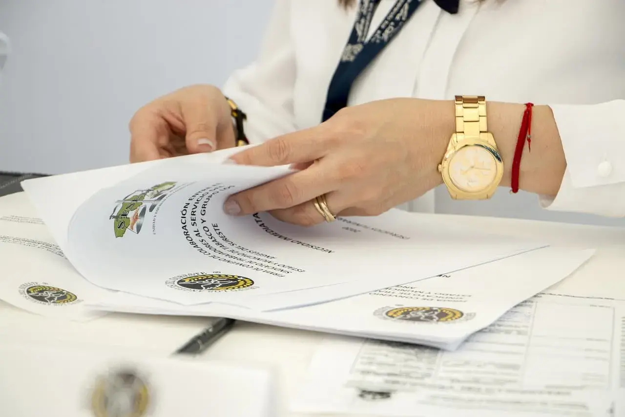 Dłonie przeglądają dokumenty, wśród których znajdują się oświadczenia o poddaniu się egzekucji. Złoty zegarek i czerwona nitka na nadgarstku.