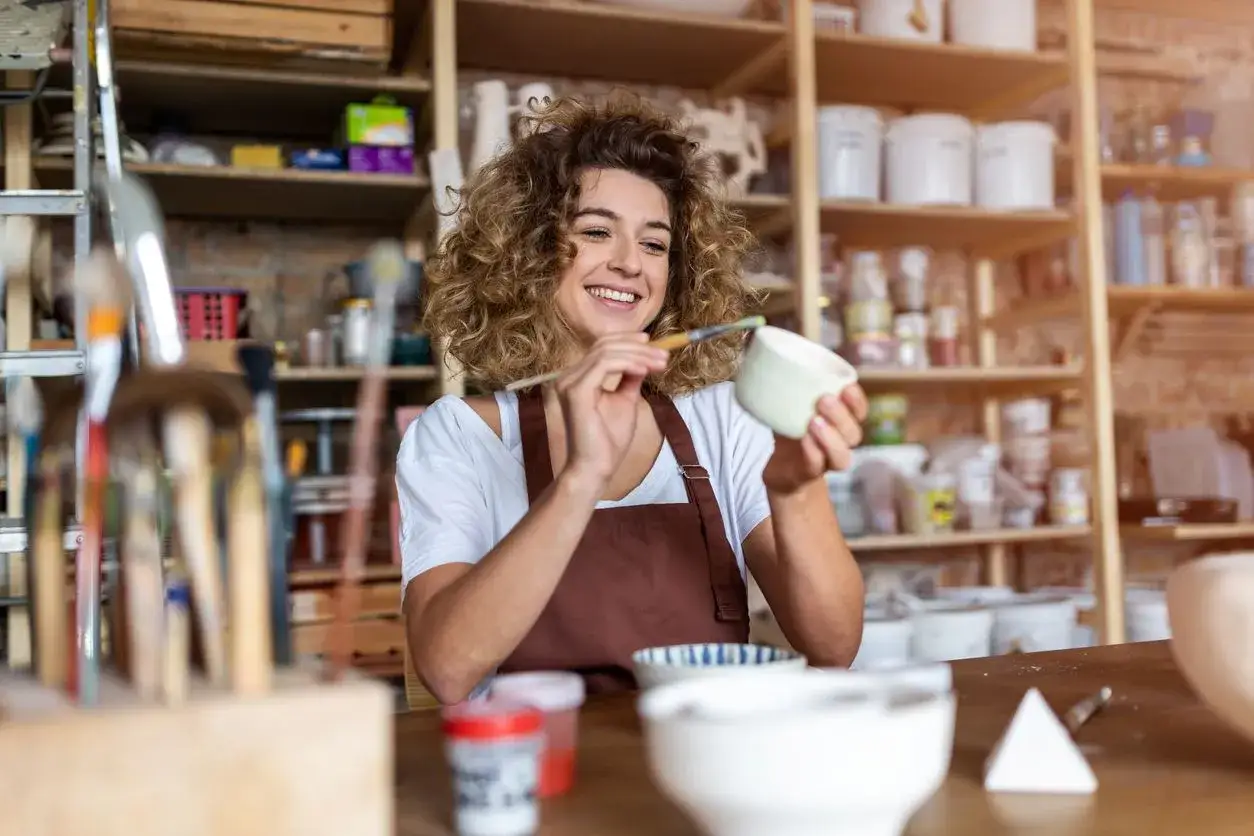 Uśmiechnięta artystka maluje ceramiczne naczynie w swojej pracowni. To jej pasja, produkcja i sprzedaż rękodzieła PKD.