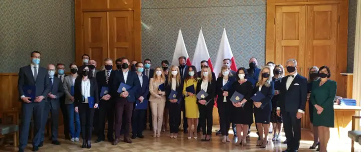 Grupa osób w garniturach i sukienkach, z flagami Polski w tle. Wśród nich radca prokuratorii generalnej, odbierający dokumenty.